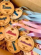 Medallas personalizadas madera fin de curso con nombres Liam, Alma y Anthonella, cintas rosa y azul
