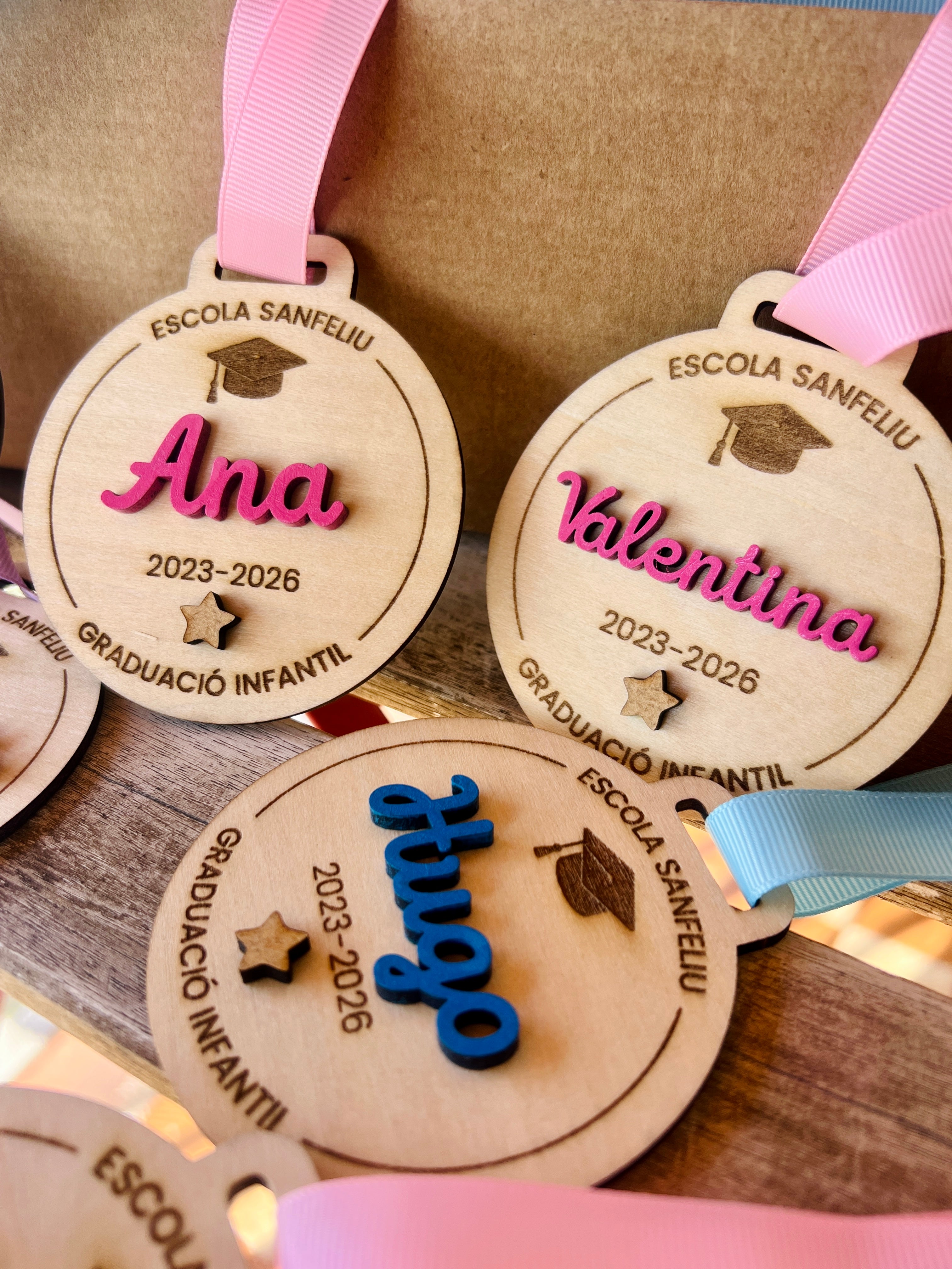 Medallas madera personalizadas con nombres Ana, Valentina y Hugo, cintas rosa y azul grosgrain